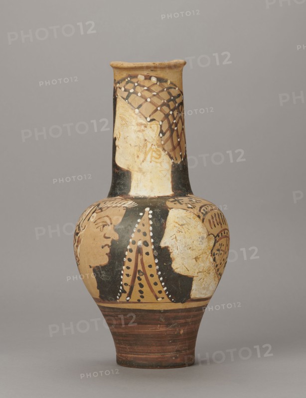 Red-figure oinochoe by the Torcop Painter, Classified Gallo-Classic Scenography; wine vessel with mythological scenes and women; 2nd half of the 4th–3rd century BC circa 300–280 BC, to be verified; Etruria place of origin; vessel; height 31.5 cm, width 16.2 cm, depth 15.6 cm, weight 0.94 kg, A painted ceramic vessel with a narrow neck and rounded body sits against a plain gray background; the vase shows a palette of tan, black, brown, white, and reddish-brown, with decorative patterns and painted human profiles around the neck and shoulder area, including a profile wearing a netlike headdress on the neck and several facing profiles on the body, dotted and lined ornamental details in white and black accenting the figures, and horizontal reddish-brown bands on the lower portion.