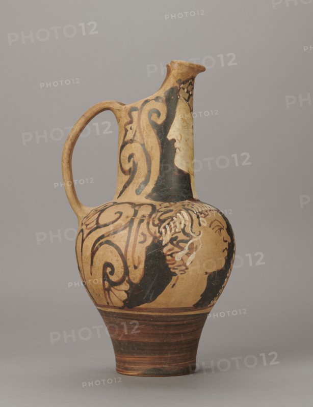 Red-figure oinochoe by the Torcop Painter, Classified Gallo-Classic Scenography; red-figure pottery, wine vessel depicting mythological scenes and women; 2nd half of the 4th–3rd century BC c. 300–280 BC, to be verified; Etruria place of origin; vessel; height 31.5 cm, width 16.2 cm, depth 15.6 cm, weight 0.94 kg, A ceramic jug with a rounded body, narrow neck, and single loop handle, displayed against a neutral gray background; the jug has an overall warm beige base color with areas of black painted decoration forming flowing scroll-like shapes and partial human profiles, and there are accents of darker brown on the lower foot and rim.