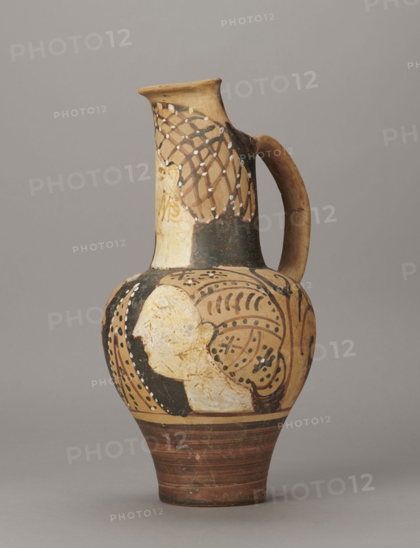 Red-figure oinochoe by the Pennsylvania Torcop Painter, Classified Gallo-Classic Scenography; red-figure pottery, wine vessel depicting mythological scenes and women; 2nd half of the 4th–3rd century BC ca. 300–280 BC, to be verified; Etruria place of origin; vessel; height 31.5 cm, width 16.2 cm, depth 15.6 cm, weight 0.94 kg., A painted ceramic jug with a rounded body, narrow neck, and single curved handle, shown against a plain gray background; the jug has an overall beige and tan base color with areas of black and dark brown paint, white painted dots and pale cream painted regions, and reddish-brown horizontal bands near the foot; decorative patterns include curved and looping lines, dotted accents, and a stylized profile-like pale shape on the body, with a netlike dark pattern on the neck and a smoothly curved handle colored tan with darkened spots.