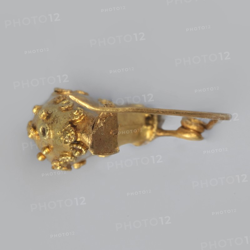 Leech fibula, brooch made of gold plate with a swollen ring decorated with filigree rings and central granulation. No sheath. 6th century BC, Etruria. Jewelry fibula. Height 1.7 cm, width 2.1 cm, depth 0.7 cm, weight 0.7 g., A small gold-colored metallic object with a textured spherical end covered in a few raised bumps and nodules, connected to a flat elongated arm and a short irregular block-like section; the object rests against a pale gray background and shows highlights and reflections in shades of bright gold and darker golden tones.
