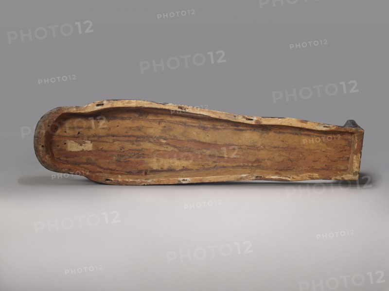 Lid of an anthropoid sarcophagus of a woman. Lid with clearly modeled legs; head sculpturally modeled with a triangular face, strong flat forward chin, large almond-shaped eyes, straight nose, and small fleshy lips. Wig is schematically designed. On the chest is a richly polychromed nine-row necklace; below it a pectoral cross in the form of a winged vulture. An offering formula runs through the center and on the sides, dividing the surface of the lid into six undecorated fields. Sacrificial formulas, vulture and necklace iconography, hieroglyphs. c. 1400–c. 1295 BC; New Kingdom, late 18th Dynasty. From Deir el-Medina grave 1352, western necropolis. Anthropoid sarcophagus; height 193 cm, width 53 cm., A long, shallow wooden object with worn, uneven edges lies horizontally against a smooth background; the wood shows a range of brown tones from light tan to deeper reddish-brown with visible grain patterns and darker streaks, and there are areas of lighter beige along the rim and patches ...