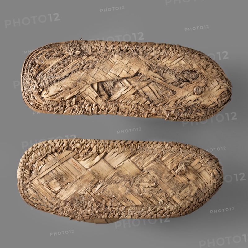 Sandals, pair of papyrus sandals made using the ribbon technique; edges finished with a cross-ribbed technique. Footwear, grave goods. c. 1550–c. 1069 BC New Kingdom, Egypt. Height 24.4 cm, width 11 cm., A pair of woven straw sandals viewed from the soles, showing a textured pattern of braided and interlaced straw fibers in various shades of tan and beige, with some darker brown strands and worn, frayed edges; the sandals rest on a smooth gray background.