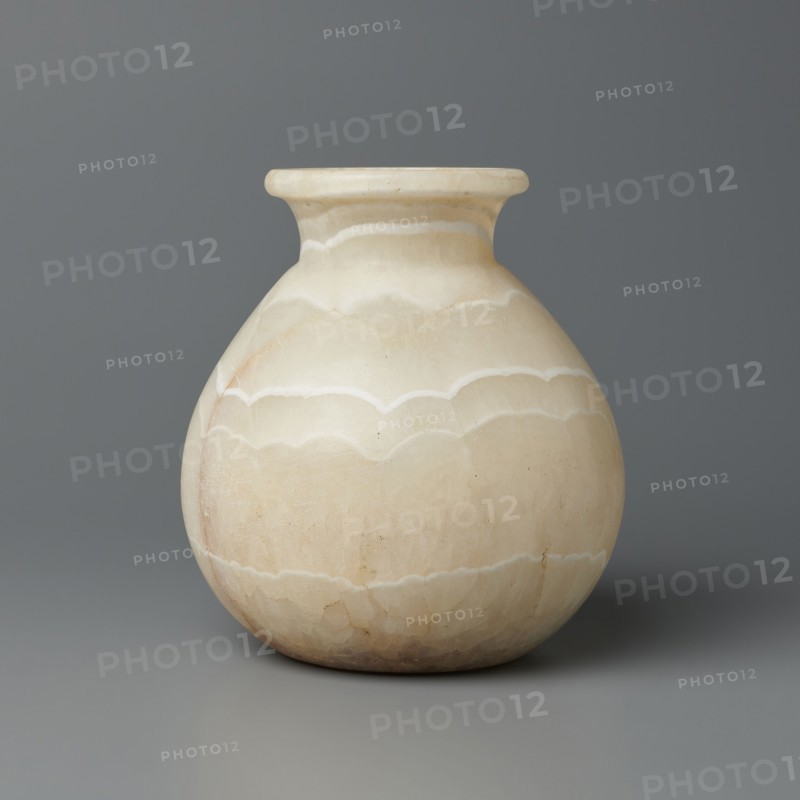 Vessel, globular vessel with a short, wide neck and a large upturned spout, flattened bottom; stone; ca. 1550–1069 BC, New Kingdom, Egypt; vessel; height 14.8 cm, width 13.4 cm, weight 1.32 kg., A rounded vase with a narrow neck and flared rim sits against a plain background; the vase is mainly creamy off-white with subtle bands and scalloped patterns in slightly darker beige and light tan tones, and there are faint natural striations and small speckles on its surface, while the background and surface it rests on are shades of gray.