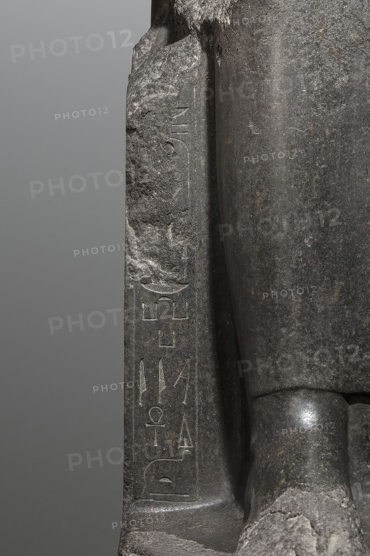 Statue of the goddess Sekhmet, Sekhmet mythology, solar discs, uraei, hieroglyphs, thrones, lionesses, Egyptian deities. Woman with a lioness's head seated on a throne, dressed in a long, close-fitting veil. Hands placed on her knees; in her left hand the goddess holds the sign of life. At the front of the throne, next to the goddess's legs, a column of hieroglyphs on each side. On the sides of the throne is carved a sema taui motif. c. 1390–1352 BC; New Kingdom, 18th Dynasty reign of Amenhotep III, found at Karnak. Sculpture; height 240 cm, width 61 cm, depth 123 cm, weight 2300 kg., A close-up view of a dark gray stone sculpture showing a section of a carved figure seated on a base, with a flat vertical slab beside the figure. The stone surface displays a range of gray tones from light to dark, with rough weathered textures and smoother polished areas. The seated figure's knee and part of the lower leg are visible, rounded and smoother with subtle highlights, while the adjacent slab ...