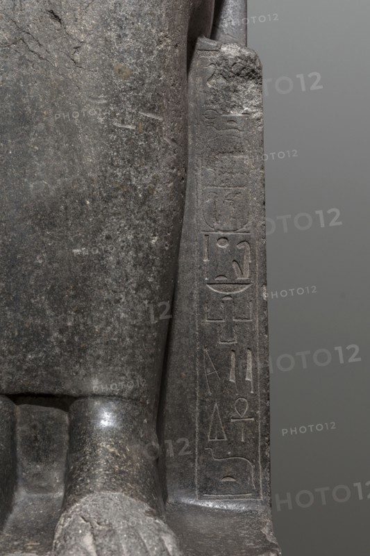 Statue of the goddess Sekhmet, Sekhmet mythology. Woman with a lioness's head seated on a throne, dressed in a long, close-fitting veil. Hands on her knees; in her left hand she holds the ankh sign of life. At the front of the throne, next to the goddess's legs, a column of hieroglyphs on each side. Sides of the throne carved with a sema-taui motif; solar discs and uraei present. c. 1390–1352 BC, New Kingdom, 18th Dynasty reign of Amenhotep III, found at Karnak. Sculpture; height 240 cm, width 61 cm, depth 123 cm, weight 2300 kg., A close-up view of a stone sculpture showing a smooth dark gray surface with lighter gray speckling and textured wear, a vertical carved panel with shallow incised markings, a rounded protruding element at the base, and a neutral medium gray background; visible colors include dark gray, light gray, and medium gray.