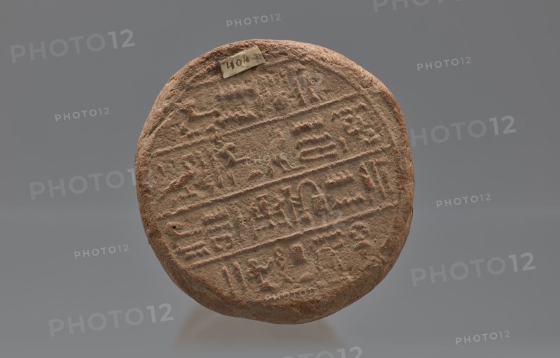 Mentuemhat's funeral cone, Mentuemhat ca. 700–650 BC. Clay cone with hieroglyphs; on the base a hieroglyphic inscription arranged in four lines. Inscriptions include name Nesiptah. Late Period, Western Thebes grave 34 on Asasipha, cone grave. Height 8.5 cm, width 12 cm, weight 0.628 kg. Davies-Macadam, Corpus No. 486, 664–332 BC., A round, flat object with a rough, textured surface in shades of brown and tan, showing a pattern of incised lines and shallow carved shapes arranged in horizontal bands across its face; the edges are irregular and slightly worn, and there is a small rectangular piece of material affixed near the top edge; the background is a smooth, neutral gray.
