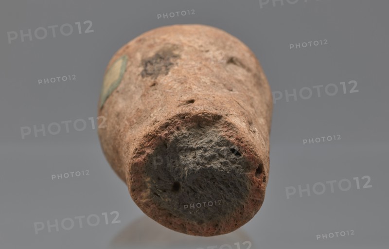 Mentuemhat's funeral cone, Mentuemhat ca. 700–650 BCE, hieroglyphs, Nesiptah ca. 700–650 BCE. On the base a hieroglyphic inscription arranged in four lines. Davies-Macadam, Corpus No. 486, 664–332 BCE; Late Period, Western Thebes grave 34 on Asasipha, cone grave. Height 8.5 cm, width 12 cm, weight 0.628 kg., A close-up view of a roughly cylindrical, weathered object with a textured, porous surface in shades of brown, reddish-brown, gray, and dark gray, shown against a smooth neutral gray background; the object's end facing the viewer appears rounded with irregular holes and rough, uneven edges, while the sides display worn, mottled patches and a small area of faded greenish color.