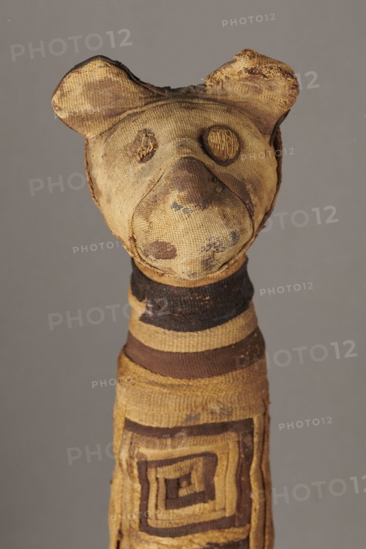 Cat mummy, Cat mummy decorated with a meander made of white and brown bandages. Materials: fabrics, linen. Subject: cats; ornament: meander. Date: 30 BC–395 AD, Roman period, Egypt. Type: mummy. Dimensions: height 27.3 cm, width 6.5 cm, depth 6 cm., A worn stuffed figure resembling an animal head and elongated body made of rough woven fabric in shades of tan, beige, brown, and dark brown, with rounded ears, a protruding snout, stitched circular eye shapes, a few darker spots and stains on the face, a banded neck with alternating dark and tan rings, and a patterned rectangular spiral design on the torso, all set against a plain gray background.