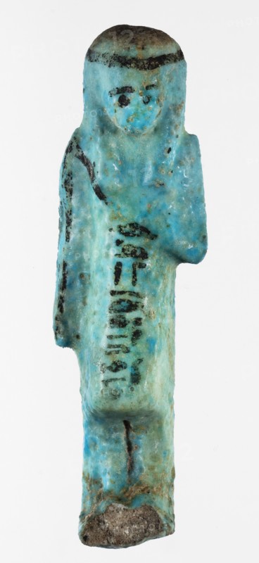 Shabti overseer Amenemone, A shabti figurine depicted in living attire, wearing a knee-length loincloth. In her left hand, bent at the elbow and folded across her chest, she holds a hoe placed on her right shoulder; the hoe's blade reaches down to her hand. The right arm hangs along the body. The face is small, triangular, with only the eyes clearly defined; other facial details are blurred. On the head is a three-part smooth wig tied with a headband, the ends of which fall down the back; the ears are uncovered. A column of hieroglyphic text on the headband at the front. Text, hoe and headband, and facial details painted in black. Circa 1069–c.945 BC, Third Intermediate Period, 21st Dynasty, Egypt. Shabti; height 8 cm, width 2.3 cm., A small statuette shaped like a standing human figure with a rounded head and stylized facial features, rendered in a turquoise-blue material with areas of darker blue and greenish patina, showing patches of black pigment on the hairline, eyes, and parts o...