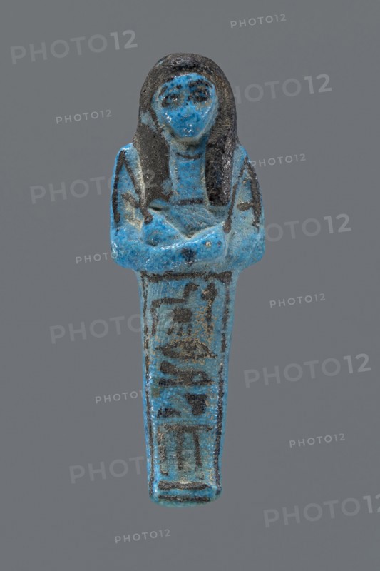 Ushabti Henut-netjeru. Mummy-shaped figurine shabti. Arms crossed on the chest, right folded over left, holding hoes in both hands; rectangular basket on the back; painted tools. Wearing a usech necklace on the chest and wide bracelets on the wrists painted jewelry. Long, smooth, three-part wig covering the ears; round face with large eyes; nose and mouth barely visible; wig and facial details painted. Painted inscription in one column on the front of the legs reading Shd Osiris Henut-netjeru, justified by the voice. Datation: ca. 1069–ca. 945 BC, Third Intermediate Period, 21st Dynasty, Egypt. Dimensions: height 9 cm, width 3 cm, depth 1.5 cm. Reference: Hnwt-ntrw -Ranke, Personennamen I, 243.25., A small blue and dark gray figurine shaped like a standing human figure with crossed arms, showing a rounded head with long dark hair and facial features, a tapered lower body with darker markings, and a textured surface with worn areas and speckles of blue and gray.