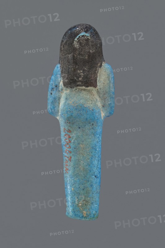 Ushabti Henut-netjeru. Mummy-shaped figurine shabti, height 9 cm, width 3 cm, depth 1.5 cm. Third Intermediate Period, 21st Dynasty, ca. 1069–ca. 945 BC. Arms crossed on the chest, right over left, holding hoes in both hands; a rectangular basket on the back with painted tools. Wearing a usech necklace and wide bracelets painted jewelry. Long, smooth three-part wig covering the ears; round face with large eyes; wig and facial details painted. Painted inscription in one column on the front of the legs reading: Shd Osiris Henut-netjeru, justified by the voice., The image shows a small human-shaped figurine viewed from the front against a plain gray background, with a rounded dark head and a long tapered body in bright blue with areas of lighter blue and speckled darker spots; the shoulders flare slightly and the surface appears textured and worn with patches of discoloration and roughness.