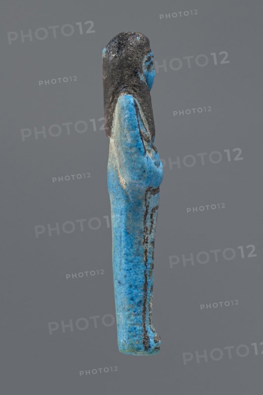 Ushabti Henut-netjeru, Henut-netjeru private name. Mummy-shaped figurine; shabti. Arms crossed on the chest, right over left, holding hoes in both hands; a rectangular basket on the back; painted tools. Wearing a usekh necklace and wide bracelets—painted jewelry. Long, smooth, three-part wig covering the ears; round face with large eyes, nose and mouth minimally modeled; wig and facial details painted. Painted inscription in one column on the front of the legs reading: Shd Osiris Henut-netjeru, justified by the voice. Date ca. 1069–ca. 945 BC, Third Intermediate Period, 21st Dynasty, Egypt. Dimensions: height 9 cm, width 3 cm, depth 1.5 cm., A small sculpted figure viewed in profile showing a standing person with long hair, arms folded against the body, and a long robe; the surface is predominantly bright blue with areas of darker blue and turquoise mottling, and the hair and some detailing appear dark gray to black, with a muted gray background.