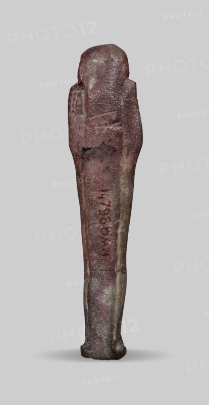 Ushabti, A shabti figurine dressed in a shroud stands on a base. On the head is a smooth wig falling in two strands on the chest. The false beard is smooth and straight. In his hands, crossed on the chest and protruding from under the shroud, he holds hoes in the shape of the letter A. 664–332 BC; Late Period 27th–30th Dynasty, Egypt. Ushabti, height 11.8 cm, width 2.8 cm., A vertically oriented slender statuette standing on a small base against a plain light gray background, with a rounded head and simplified body shape; the surface appears rough and weathered with visible texture variations and some cracks, and the overall color palette includes dark reddish-brown, muted brown, and areas of grayish-beige, with a few darker gray patches and subtle lighter speckles.