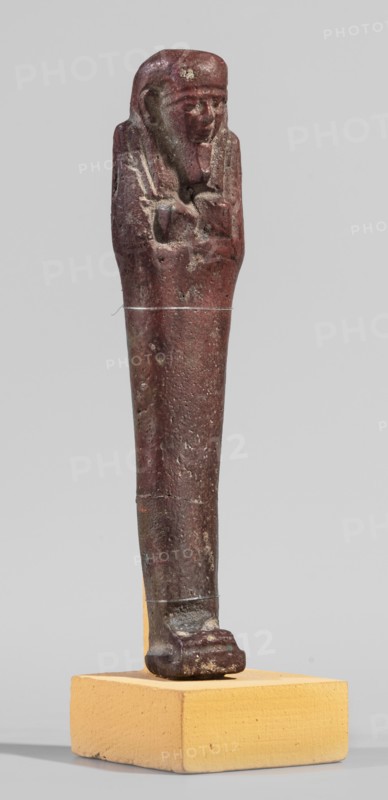 Ushabti, A shabti figurine dressed in a shroud stands on a base. On the head is a smooth wig falling in two strands on the chest. The false beard is smooth and straight. In his hands, crossed on the chest and protruding from under the shroud, he holds hoes in the shape of the letter A. 664–332 BC; Late Period 27th–30th Dynasty, Egypt. Ushabti; height 11.8 cm, width 2.8 cm., A small statuette of a standing figure with folded arms displayed on a rectangular block base; the figure has a rounded head with visible facial features and a headdress, and the body tapers down to a flat foot resting on the base; the surface shows an aged, rough texture with worn areas and small pits; colors visible include deep reddish-brown on the figure, lighter brown and pale tan on the base, and small areas of lighter discoloration on the figure.