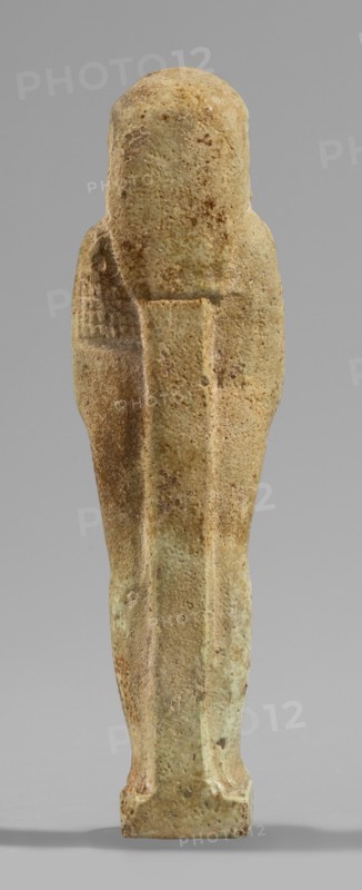 Ushabti, Wigs, beards facial hair, funerary figurine mummy-shaped, hoes, grave goods. Standing on a base, supported at the back by a pillar/pilaster. Arms crossed on the chest, only the hands protrude from under the shroud, placed at the same level slightly apart. Hoes in both hands; the ends of the hoes' handles are slightly curled. In the right hand an additional twisted rope, on which a basket is slung over the left shoulder. Slightly convex tools. On the head a three-part, smooth wig. A long, straight beard tapering toward the bottom, braided into a plait. Large ears, almond-shaped eyes, pronounced eyebrows, broad nose, full lips in a slight grimace. Not inscribed. 664–332 BC; Late Period 26–30th Dynasty, Egypt. Shabti; height 11.7 cm, width 3.2 cm., A vertical stone or ceramic figurine viewed from the back, with a rounded top and a narrow rectangular base, showing a tapered, elongated body with subtle raised ridges along the center and sides, a pronounced seam down the middle of t...