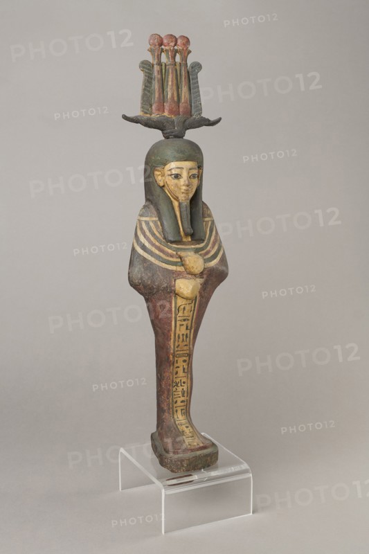Osiris, Osiris mythology. Figurine in the form of a human mummy wearing a hemhem crown with ram's horns. On the chest is a usech necklace with falcon heads at the ends. Clenched hands are placed on the chest, one above the other. Below the right hand begins a column of text with the name and filiation. At the back, from the edge of the wig to the feet, a pillar with a column of hieroglyphic inscriptions — the beginning of a hymn to Osiris, the heir of Atum. The figurine is painted cherry red, the visible parts of the body are yellow, the wig is navy blue, and the necklace consists of white, red, and blue stripes. The background of the inscriptions is yellow. The figurine belongs to Isetemachbit, a musician of the god Amun-Re, daughter of the priest Mentuemhat; her mother was probably Takereret, known as Heres. Similar to type IV B according to Raven. 4th–3rd century BC; 30th Dynasty–early Ptolemaic period, Egypt. Sculpture; height 54.5 cm, width 12.5 cm, depth 9.5 cm., A painted wooden...