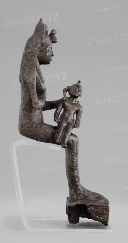 Figurine of the goddess Isis nursing Horus. The figurine depicts a seated Isis with the child Horus on her lap; Isis supports Horus with her left hand and offers him her breast with her right. Isis is dressed in a narrow, close-fitting robe and wears a fragment of the base of a crown on her head. Horus is naked, wears a neck ornament, has a lock of youth hanging from the right side of his head, and a uraeus placed above his forehead. A hieroglyphic votive text is inscribed on the base. Late Period, 525–343 BC 27th–30th Dynasty, Egypt. Sculpture; height 21.7 cm, width 7 cm, depth 10.5 cm, weight 1.22 kg., A dark brown metallic sculptural figure seated on a clear support, shown in profile, holding a smaller seated child figure on its knee; the larger figure has a textured surface, an elongated headdress with a rounded protrusion at the forehead, a visible eye and nose in profile, and an arm extended to support the child; the child figure is also dark brown metallic with a small headdress...