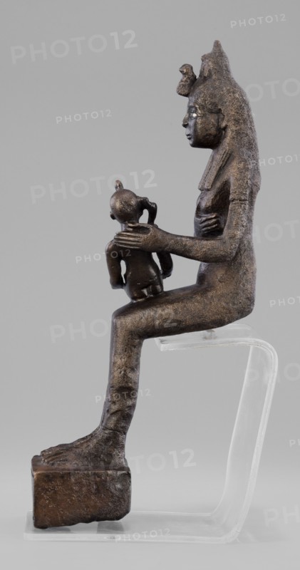 Figurine of the goddess Isis nursing Horus. The figurine depicts a seated Isis with the child Horus on her lap. Isis supports Horus with her left hand and offers him her breast with her right. Isis is dressed in a narrow, close-fitting robe. On her head is a fragment of the base of a crown. Horus is naked, wearing a neck ornament; his head bears a lock of youth hanging from the right side and a uraeus placed above his forehead. On the base of the figurine is a hieroglyphic votive text. Late Period, 525–343 BC 27th–30th Dynasty, Egypt. Sculpture. Height 21.7 cm; width 7 cm; depth 10.5 cm; weight 1.22 kg., A dark brown metallic sculpture of a seated adult figure in profile holding a smaller childlike figure on its knee, both figures showing textured surfaces and defined facial profiles, the adult wearing a tall headdress and a long garment draped along the back, the adult's hand resting on the child's shoulder while the child faces forward with a rounded head and small limbs, the adult s...