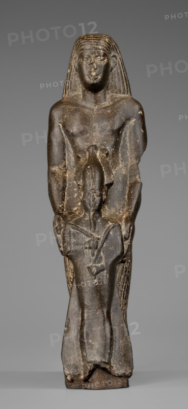 Figurine of a priest with a statuette of the god Osiris. The priest is shown standing, holding in front of himself with both arms a statuette of Osiris, which reaches below his waist. The priest is dressed in a long, pleated loincloth. On his head he wears a wig pushed halfway down his forehead and reaching to his shoulders. The musculature of the torso and arms is clearly defined, with a marked vertical line dividing the torso, and with strongly modeled collarbones. The priest's face is small, modern, with slightly convex, smooth cheeks. Slanted eyes, eyebrows with a gentle arch, artistically rendered. Large ears. Osiris is depicted in a shroud, wearing an atef crown, the front of which is decorated with a uraeus. In his right hand he holds a Nechacha whip, in his left a Heka scepter. The face is round, with large eyes and a slightly smiling mouth. The priest's figure is leaning against a rear pillar reaching to his wig, on which a hieroglyphic inscription is carved; some of the chara...