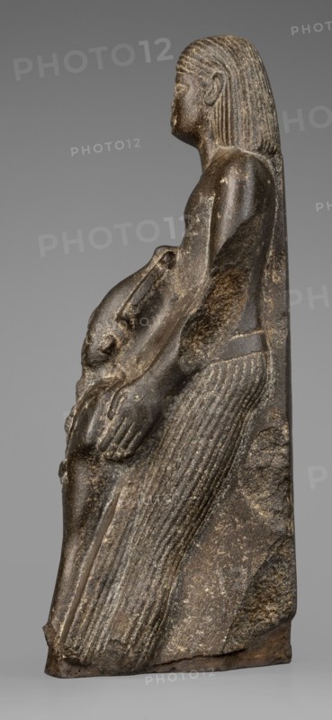 Figurine of a priest with a statuette of the god Osiris, The priest is shown standing, holding in front of himself with both arms a statuette of Osiris, which reaches below his waist. The priest is dressed in a long, pleated loincloth. On his head he wears a wig pushed halfway down his forehead and reaching to his shoulders. The musculature of the torso and arms is clearly defined, with a marked vertical line dividing the torso, and with strongly modeled collarbones. The priest's face is small, modern, with slightly convex, smooth cheeks. Slanted eyes, eyebrows with a gentle arch, artistically rendered. Large ears. Osiris is depicted in a shroud, wearing an atef crown, the front of which is decorated with a uraeus. In his right hand he holds a Nechacha whip, in his left a Heka scepter. The face is round, with large eyes and a slightly smiling mouth. The priest's figure is leaning against a rear pillar reaching to his wig, on which a hieroglyphic inscription is carved; some of the chara...