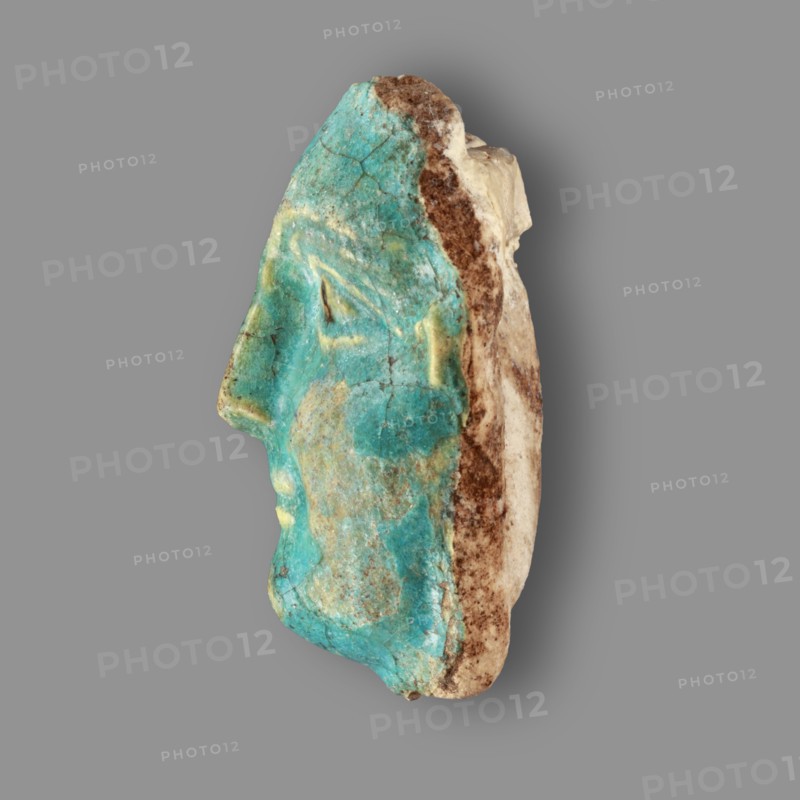Fragment of a figurine. Oval male face with a protruding chin, small mouth, straight nose, and almond-shaped eyes. Small, protruding ears; forehead receding backward. Unfinished at the back. Egyptian, Ptolemaic Period, 332–30 BC. Figurine, height 5.9 cm, width 4.5 cm, depth 2.5 cm., A side view of a sculpted human face fragment with a turquoise-green glazed surface showing weathered and cracked areas, a few pale yellow highlights on the forehead and near the mouth, and an exposed rough beige and brown stone backing along the rear edge, all set against a uniform gray background.