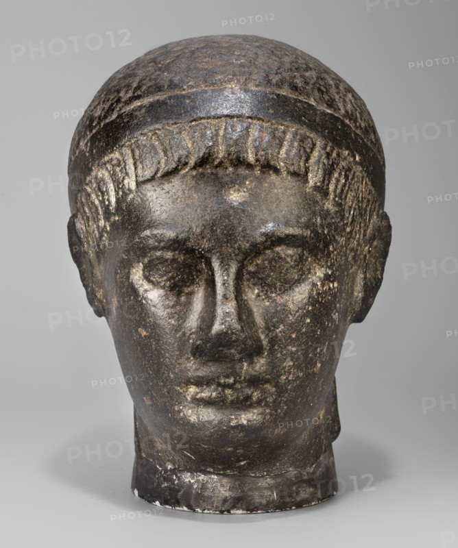 Head of a Ptolemaic prince, unknown creator; diadems; Ptolemy XV Caesarion ruler of Egypt; 47–30 BC probably; kings; Egyptian sculpture; Egyptian art; hairstyles. Head of a man with short hair tied with a headband crossed at the nape of the neck and falling at the ends to the neck. Short, wide strands of hair protrude from under the headband onto the forehead. The face is small, with regular features, a straight nose widening significantly downward, prominent lips, a clearly emphasized chin, large deep-set eyes, prominent eyebrows, small ears close-fitting to the head. The upper fragment of the pilaster is visible at the back, with the head resting on it. With underlay 20.5, 44–30 BC; Ptolemaic period end of the reigns of Caesarion and Cleopatra, Egypt; sculpture. Dimensions: height 19.3 cm, width 13 cm, depth 17 cm; weight 7.532 kg., A sculpted head made of dark stone with lighter worn areas, showing a smooth rounded top, a band around the forehead, and short stylized hair along the h...