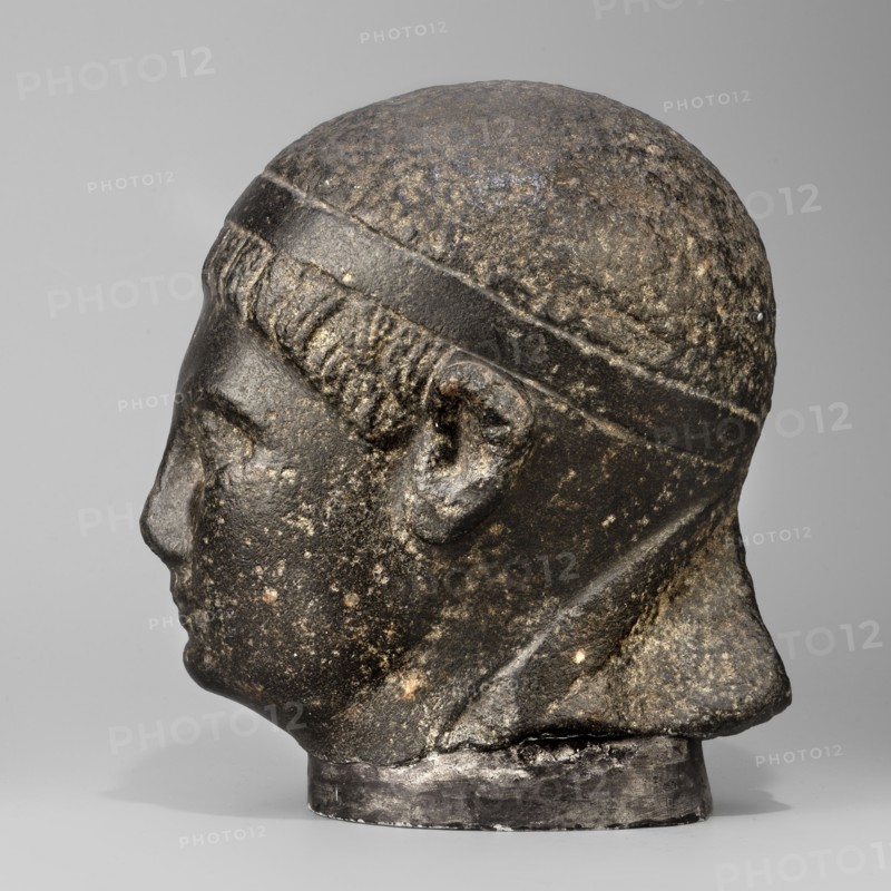 Head of a Ptolemaic prince; diadems; Ptolemy XV Caesarion ruler of Egypt; 47–30 BC probably; Egyptian sculpture, Ptolemaic period end of the reigns of Caesarion and Cleopatra, 20.5, 44–30 BC; sculpture of a man with short hair tied with a headband crossed at the nape of the neck and falling at the ends to the neck. Short, wide strands of hair protrude from under the headband onto the forehead. The face is small, with regular features, a straight nose widening significantly downward, prominent lips, a clearly emphasized chin, large deep-set eyes, prominent eyebrows, small ears close-fitting to the head. The upper fragment of the pilaster is visible at the back, with the head resting on it. Material: Egyptian sculpture. Dimensions: height 19.3 cm, width 13 cm, depth 17 cm; weight 7.532 kg., A close-up view of a sculpted head in profile showing the left side of the face, ear, and back of the head. The surface appears rough and weathered with textured areas across the scalp and face. The h...