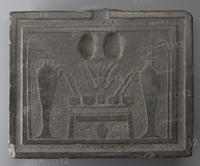 Offering table with carved offerings;, Offering table with offerings depicted: bread and two slender vessels, behind which lotus flowers are visible on high stems. Iconography: offerings, grave goods. Ptolemaic Period, 332–30 BC, Egypt. Dimensions: height 8.5 cm; width 36 cm; depth 29 cm; weight 23.444 kg., A rectangular carved stone plaque in shades of gray with a raised border contains a recessed central panel showing a symmetrical arrangement: a low table or platform with a row of rounded objects on top, flanked by two tall slender vase-like forms with curved handles or spouts, stylized plant motifs rising behind the table, and two oval recessed shapes above; the surface shows a textured, slightly pitted finish and worn edges.