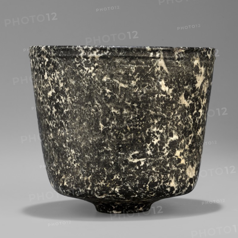 Stone vessel, Königsberg provenance, repossession Frombork; Warmian-Masurian Voivodeship, stone vessel, ritual vessel. Vessel with a quadrangular bottom and an almost cylindrical body profiled below the rim. The vessel is supported by a small, round foot. 332–30 BC; Ptolemaic Period One should also consider the possibility that this vessel is not very early. J. Velvet, Egypt, vessel, height 8.8 cm, width 10 cm, weight 0.63 kg, A close-up view of a rounded cup or vessel with a slightly flared rim, showing a mottled surface of dark gray and off-white patterns that create a marbled appearance; the vessel has a short, slightly raised foot and casts a soft shadow on a smooth gray background.
