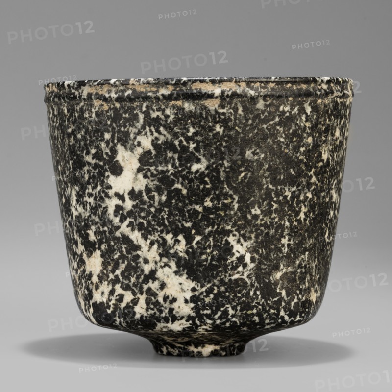 Stone vessel, Königsberg provenance, repossession Frombork; Warmian-Masurian Voivodeship. Stone ritual vessel with a quadrangular bottom and an almost cylindrical body profiled below the rim, supported by a small round foot. 332–30 BC; Ptolemaic Period possibility it is not very early — J. Velvet. Egypt; vessel. Height 8.8 cm, width 10 cm, weight 0.63 kg., A cylindrical cup with a slightly flared rim and a small rounded foot sits against a neutral backdrop; its surface shows a speckled marbled pattern of black and white with a few beige spots, the rim area displaying some worn beige and off-white along the edge, and a soft shadow beneath the cup.