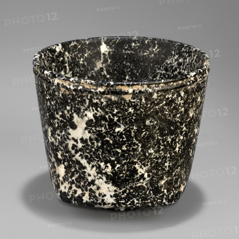 Stone vessel, Königsberg provenance, repossession Frombork; Warmian-Masurian Voivodeship. Stone vessel, ritual vessel. Vessel with a quadrangular bottom and an almost cylindrical body profiled below the rim. The vessel is supported by a small, round foot. 332–30 BC; Ptolemaic Period one should also consider the possibility that this vessel is not very early. Egypt, vessel. Height 8.8 cm, width 10 cm, weight 0.63 kg., A close-up of a rounded cup-like object with a mottled black and white speckled surface, showing a slightly flared rim with a narrow raised band near the top, smooth interior with the same speckled pattern, subtle wear and small lighter patches on the exterior, and a soft gray background casting a faint shadow beneath the object; the visible colors include black, white, gray, and small areas of tan.