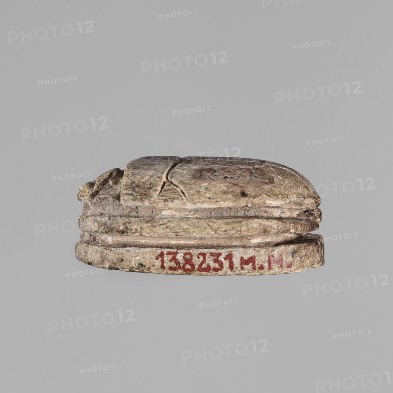 Scarab with a silhouette of a ruler on a throne. The scarab has on its base a representation of a royal figure sitting on a throne, with a scepter and a whip in its hands, possibly Osiris, and a second, smaller standing figure. ca. 1550–1295 BC; New Kingdom, 18th Dynasty, Egypt. Scarab amulet; height 3.1 cm, width 2.2 cm., A small carved object shaped like a rounded scarab rests against a plain light gray background; the object has a weathered surface in shades of beige, tan, and light brown with darker brown spots and subtle greenish-gray areas, and it shows a few visible cracks and worn edges; the sides feature layered ridges and a few areas of darker discoloration, and the overall texture appears rough and aged.