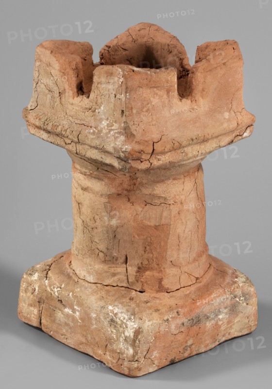 Horned altar serving as a censer, ceremonial vessel/altar with square base and cylindrical shaft, lightly profiled Doric base and capital topped with vertical walls with a single rectangular cutout on each side. Ptolemaic Period, 332–30 BC Tell Atrib, altar; height 14.5 cm, width 8 cm, weight 0.454 kg, A weathered terracotta object in shades of reddish-brown, tan, and beige sits against a neutral gray background, showing a rough, cracked surface with areas of lighter whitish residue; its form includes a square base transitioning into a cylindrical midsection and topped by an irregular, crown-like upper portion with deep indentations and a hollow center, exhibiting visible chip marks, fractures, and textured, uneven edges.