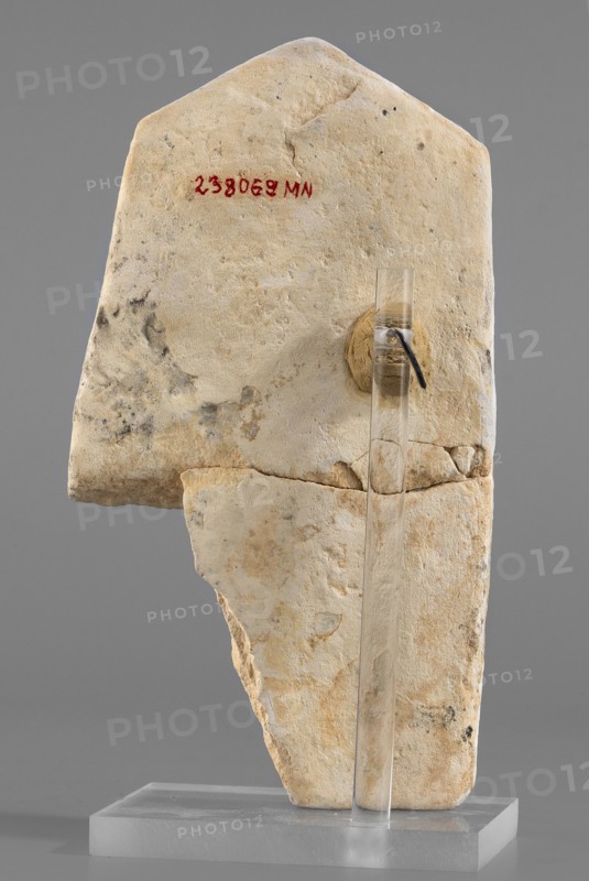 Naos stele with a female figure, Small naos-shaped stele, slightly tapered at the top and triangular; naos design marked with a flat engraving. A female figure is schematically represented inside. Egyptian deities, religious rituals, architectural motifs. Late Period–Ptolemaic Period, 664–30 BC. Findspot: Tell Atrib. Stele; height 12 cm, width 6.5 cm., A pale beige stone fragment with rough, weathered surfaces and irregular edges is shown standing upright on a transparent rectangular support; the fragment has areas of darker gray and brown discoloration and mottling across its surface, a circular patched area of slightly darker beige where the support is attached, and a clear vertical rod fixed to the front of the piece; the background is a smooth mid-gray.