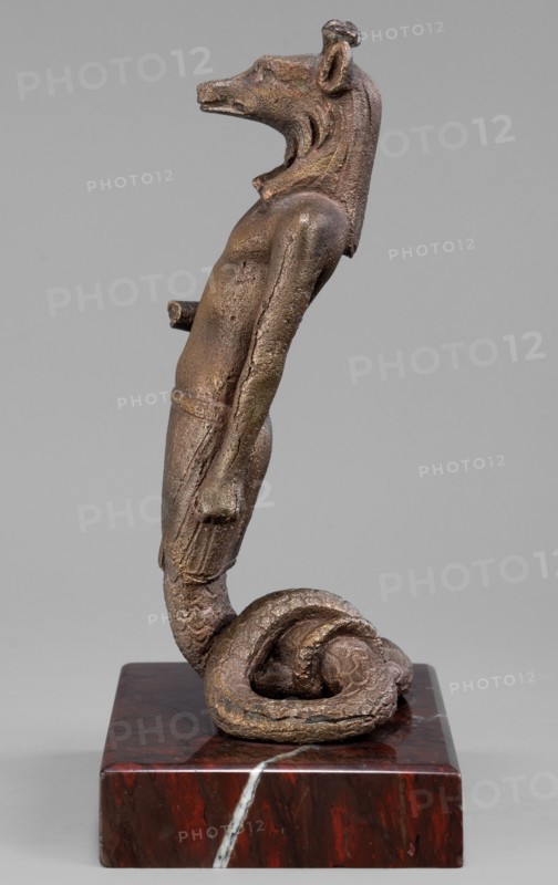 The god Anubis in the form of a jackal-headed deity; Anubis mythology. Figurine of Anubis in the form of a human-jackal-serpent in an upright posture, torso slightly tilted back, with a jackal head with a strongly elongated muzzle and a mane flowing down the back. Dressed only in an Egyptian apron, below which the torso merges into the body of a snake with a striated abdomen and scaled skin, curled in a horizontal figure eight that supports the figurine. Left arm held straight alongside the body with a clenched fist; right arm bent at the elbow with the forearm extended forward. Three protrusions on the head between the ears. Late 1st–2nd century AD, Roman period, Egypt. Terracotta/bronze? figurine. Height 13.5 cm, width 7.1 cm, weight 0.936 kg with base., A bronze-colored sculpture of a humanoid figure with an animal-like head stands on a rectangular dark red and black marble base with lighter veining; the figure has a textured surface with areas of brown, bronze, and greenish patina,...