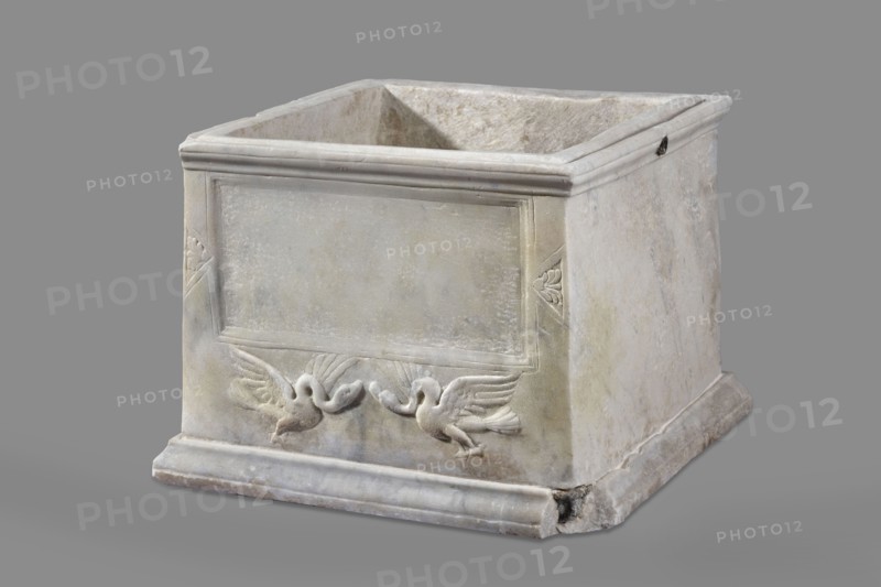 Urn with a depiction of swans. Main plate: the urn's box is trapezoidal, tapering toward the top, placed on a profiled base. The front surface is sparsely decorated; the main area features a tabula ansata whose ears are decorated with palmettes. Below the inscription field are two swans in deep relief, facing each other with strongly arched necks and appearing to take flight, their outstretched wings touching the inscription frame. The upper part is crowned by a profiled cornice; on the inner upper edge a rectangular protrusion for securing the lid is visible. Side walls: no decoration; metal pins visible under the cornice on both sides. Lid: original missing. Dated to the 2nd half of the 1st century AD probably 3rd quarter of the 1st century AD based on analogies; broadly 2nd half of 1st–1st half of 2nd century AD. Roman Empire Rome. Sepulchral funerary urn, weight 45.5 kg. Maker: unknown Roman workshop., A square stone box with an open top, showing worn and slightly rough surfaces in...