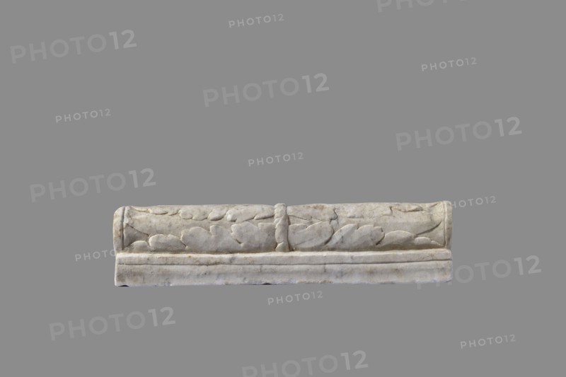 Urn with a depiction of swans, unknown Roman workshop. Urn with sepulchral funerary function, Roman, dated to the 2nd half of the 1st century AD probably 3rd quarter of the 1st century AD; Roman Empire 27 BC–476, findplace: Rome. Weight 45.5 kg. Main plate: The urn's box is trapezoidal, tapering toward the top, placed on a profiled base. The front decoration is sparse; the main surface is occupied by a tabula ansata whose ears are decorated with palmettes. Below the inscription field are two swans in deep relief, facing each other with strongly arched necks, appearing to take flight with outstretched wings touching the inscription frame. The upper part is crowned by a profiled cornice. On the inner upper edge a rectangular protrusion for securing the lid is visible. Side walls: no decoration. Metal pins visible under the cornice on both sides. Lid: original missing., A horizontal stone object with carved rounded leaf-like shapes along its front surface, showing a central raised vertica...