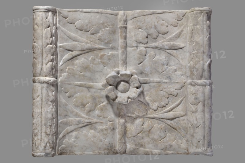 Urn with a depiction of swans, unknown Roman workshop. Sepulchral funerary urn, Roman, dated to the 2nd half of the 1st century AD probably 3rd quarter of the 1st century AD — period: Roman Empire 27 BC–476. Main plate: trapezoidal box tapering toward the top, placed on a profiled base. Front decorated sparsely; main surface occupied by a tabula ansata with palmette-decorated ears. Below the inscription field are two swans in deep relief, facing each other with strongly arched necks, appearing to take flight with outstretched wings touching the inscription frame. Upper part crowned by a profiled cornice; rectangular protrusion on inner upper edge to secure a lid. Side walls: undecorated. Metal pins visible under the cornice on both sides. Lid: original missing. Findspot: Rome. Weight: 45.5 kg., The image shows a rectangular carved stone panel with a central raised floral rosette and surrounding carved leaf motifs; the stone is predominantly off-white to light gray with areas of darker ...