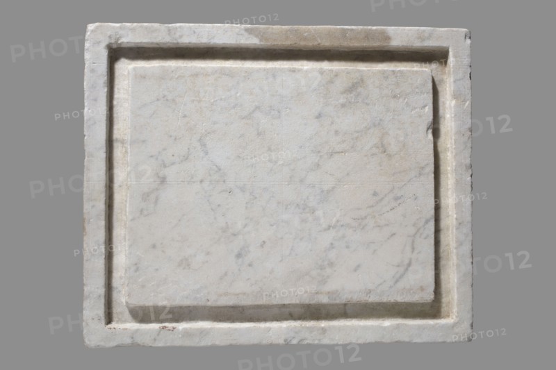 Urn with a depiction of swans, unknown Roman workshop. Main plate: the urn's box is trapezoidal, tapering toward the top and placed on a profiled base. The front is sparsely decorated; the main surface is occupied by a tabula ansata whose ears are decorated with palmettes. Below the inscription field are two swans in deep relief, facing each other with strongly arched necks and outstretched wings touching the inscription frame. The upper part is crowned by a profiled cornice; on the inner upper edge a rectangular protrusion for securing the lid is visible. Side walls: no decoration; metal pins visible under the cornice on both sides. Lid: original missing. Dated to the 2nd half of the 1st century AD probably 3rd quarter of the 1st century AD — Roman Empire 27 BC–476. Place of origin/find: Rome. Urn Roman. Weight: 45.5 kg., A square slab with a recessed rectangular panel fills the image, showing a surface of white and light gray marble with subtle veining and faint mottling, surrounded ...