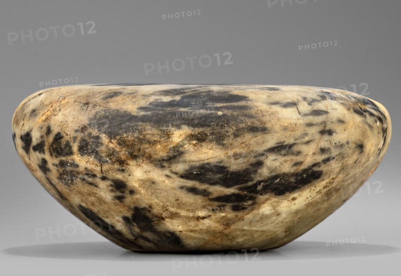 Stone vessel, stone, in the shape of a bird's nest with flat broad shoulders and a small unbordered spout opening diameter 6.1 mm, walls narrowing in an arch toward a flat undivided foot; ca. 2890–2613 BC, Early Dynastic Period, 2nd–3rd Dynasty, Egypt; vessel; height 6.5 cm, width 15.3 cm, weight 1.0 kg., A smooth, rounded stone bowl with a low, wide profile sits against a plain background; its surface shows a marbled pattern of creamy beige and light tan mixed with patches and streaks of dark gray and black, with subtle areas of brownish discoloration and faint hairline cracks, and the lighting creates soft highlights and gentle shadows along its curved contours.