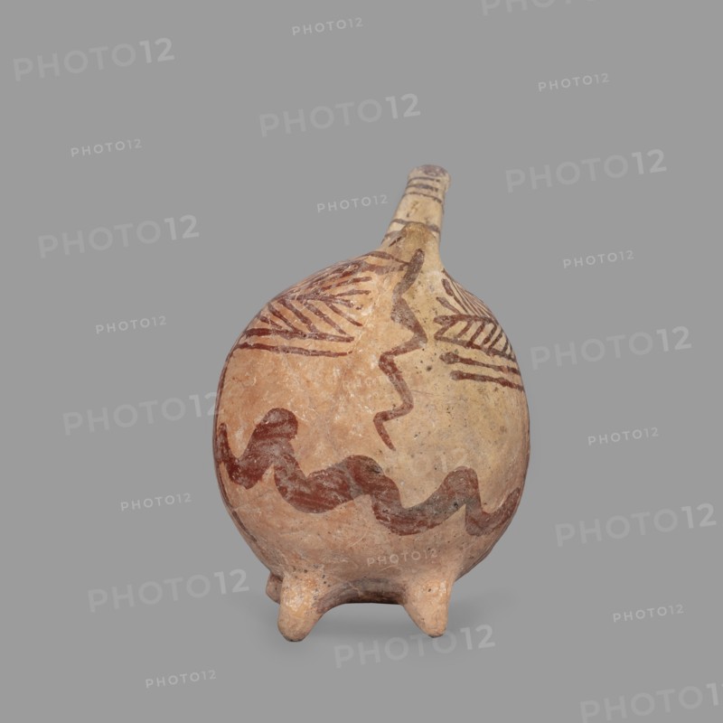 Vessel in the shape of an animal, unknown Cypriot workshop. Fantastic animal-shaped vessel with a long thin neck ending in a triangular, slightly flattened head. Eyes marked by depressions; oblong flat ears. Geometric decoration covers the entire surface: at the widest point a band of zigzag filled with a checkered pattern; herringbone stripes radiate at right angles front and back from this band; beneath these are wavy paint stripes. Series of thin lines on the neck and a hole in the front under the neck. Late Bronze Age II, 1900–1600 BC, Cyprus. Vessel; height 10.7 cm, width 12.5 cm, depth 6.8 cm., A rounded ceramic vessel with a narrow spout and a few short rounded feet, shown against a plain gray background. The vessel surface is mainly tan and light beige with areas of warm reddish-brown. There are several painted wavy and zigzag bands in a darker brown-red color encircling the body and extending toward the neck, along with a few linear hatched motifs near the shoulder. The spout ...