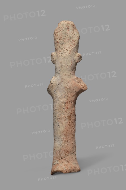 Votive figurine, Cyprus; male figurine with tiara triple crown, votive offering. Figurine of a man in a high headdress, with a cylindrical body and cylindrical arms raised upwards and outstretched to the sides. The face, neck, and torso are of equal thickness; only a large nose is visible. Large ears are attached to the sides of the head. Dated 750–600 BC formerly 7th–6th century BC. Terracotta figurine, height 14.2 cm, width 5.0 cm., A tall, slender, upright textured object resembling a rough sculpture with an elongated body and a slightly wider top that has two small protrusions on its sides, standing against a plain gray background; the surface shows a mix of beige, light brown, reddish-brown, and small flecks of darker gray, with uneven, pitted texture and some smoother worn areas.