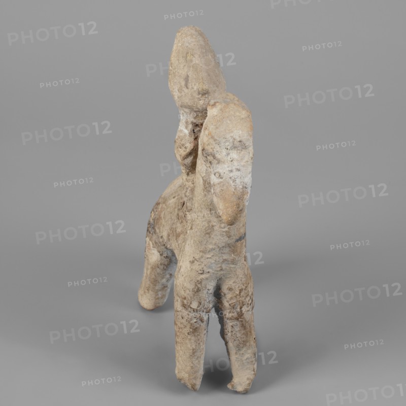 Figurine of a rider on a horse, Cyprus. The entire piece is schematic, the overall shape is emphasized and lacks fine detail. The horse is small with a short torso, cylindrical slightly tapering legs, and an unmarked tail. The neck is very long and vertical; the muzzle is large; the eyes are slightly convex; the ears are small; the mane is short and trimmed. The rider sits upright, close to the horse’s neck; the torso is short; the legs and arms are indicated. The head is large with a clearly defined profile and a particularly prominent nose; the eyes are unmarked. A vertically sloping adjacent ridge at the back of the head may indicate a helmet plume, though the helmet itself is not marked, only an overdeveloped top of the head suggests its presence. Dated 750–600 BC previously dated 7th–6th century BC. Figurine; height 14.9 cm, width 11.1 cm, depth 4 cm., A small weathered sculpture of a figure mounted on an animal, shown against a plain gray background, with the sculpture displaying...