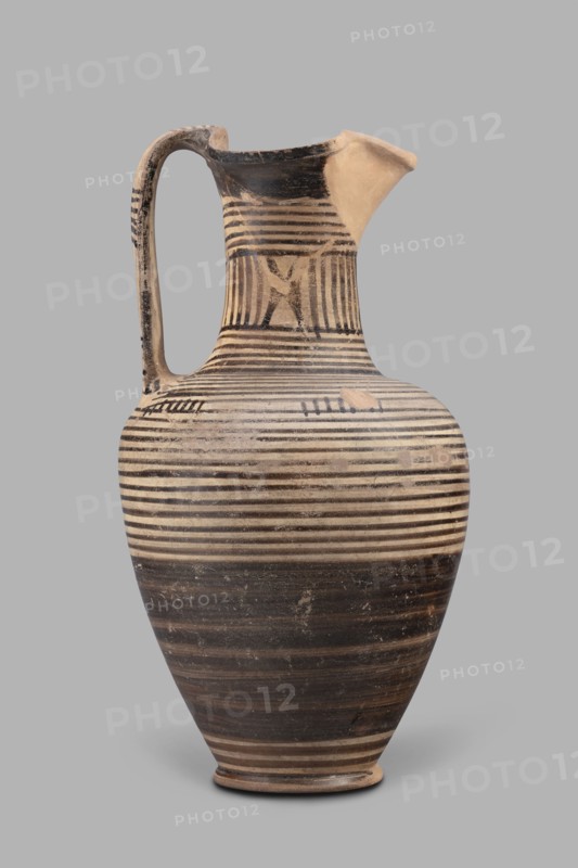 Oinochoe, Geometric oinochoe with a low foot, a gradually widening belly, distinct shoulders, a slender neck, and a tripartite rim characteristic of this vessel form. The vase runs from the shoulder to the rim. Halfway up the belly it is covered with black finish; the rest of the vessel is decorated with closely arranged parallel horizontal lines encircling the entire vessel. The central part of the neck is decorated with vertical stripes. Oinochoe was used for drawing and pouring wine Greek: oinos - wine and is a typical Greek vessel. Southern Italy Cumae workshop, end of the 8th century BC also dated end of 8th–beginning of 7th century BC; described as an Italian imitation of the Proto-Corinthian workshop. Vessel oinochoe; weight 0.476 kg; height 23.2 cm; diameter 12.2 cm., A tall ceramic jug with a rounded body, narrow neck, and spout, featuring a single curved handle; the surface shows a pattern of horizontal bands and stripes in black and beige, with a few areas of worn or lighter...
