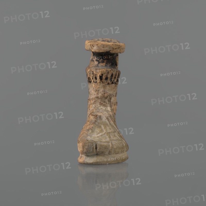 Foot-shaped aryballos, Ionian workshop, aryballos, antique vessel. Yellow-orange clay with mica; matte black engobe on the surface. Shaped as a foot shod in a sandal with a thick sole connected to braided leather straps. The foot ends above the ankle and merges into a narrow neck terminating in an outward-turned rim, decorated from above with radiating lines. A wide, ribbon-like handle is attached vertically to the spout, bearing an image of a rooster surrounded by dots. Preservation: the front part of the foot is filled with plaster. Date: end of the 6th – beginning of the 5th century BC. Findspot: Ionia, Turkey/Greece., A small carved object resembling a stylized foot or shoe stands upright against a plain gray background, showing a weathered surface with a mix of beige, tan, brown, and darker blackened areas; the sole and toe area are rounded and display worn texture and subtle carved lines, the midsection tapers upward with irregular rough patches and lighter beige highlights, and ...