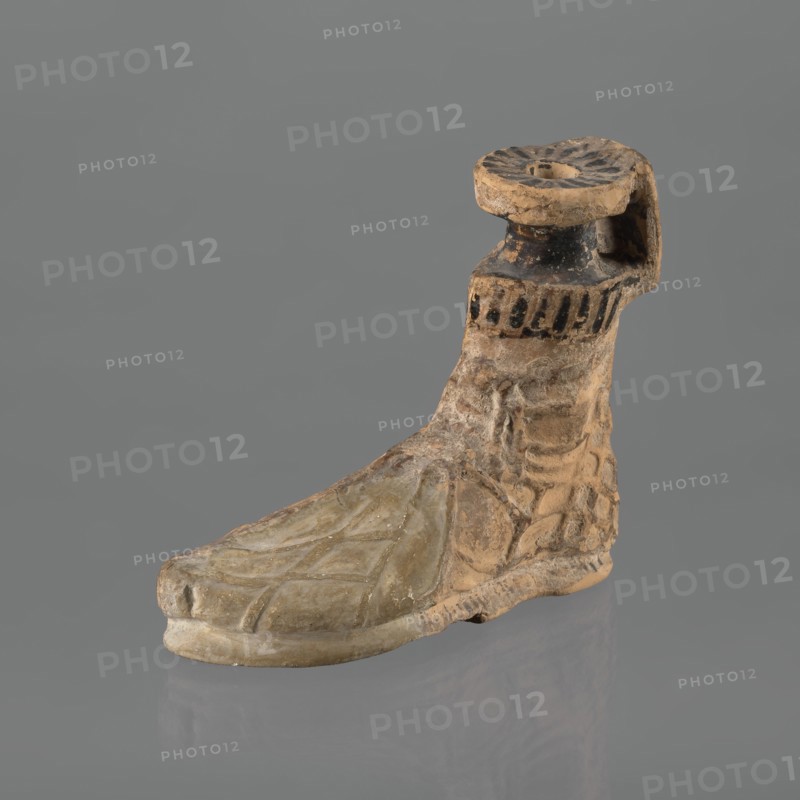 Foot-shaped aryballos, aryballos, antique vessel; Yellow-orange clay with mica; matte black engobe on the surface. Shaped as a foot wearing a sandal with a thick sole connected to braided leather straps. The foot ends above the ankle and merges into a narrow neck terminating in an outward-turned rim, decorated from above with radiating lines. A wide, ribbon-like handle is attached vertically to the spout, on which there is an image of a rooster surrounded by dots. Preservation condition: the front part of the foot is filled with plaster. End of the 6th–beginning of the 5th century BC; unknown Ionian workshop; Ionia Turkey/Greece., An object shaped like a foot with an attached ankle and short calf, rendered in earthy tan and beige tones with areas of darker brown and black, shows a textured surface with carved lines suggesting toes and instep on the forepart and layered, irregular surface on the ankle and heel; the top of the calf has a circular, dish-like element with radiating grooves...