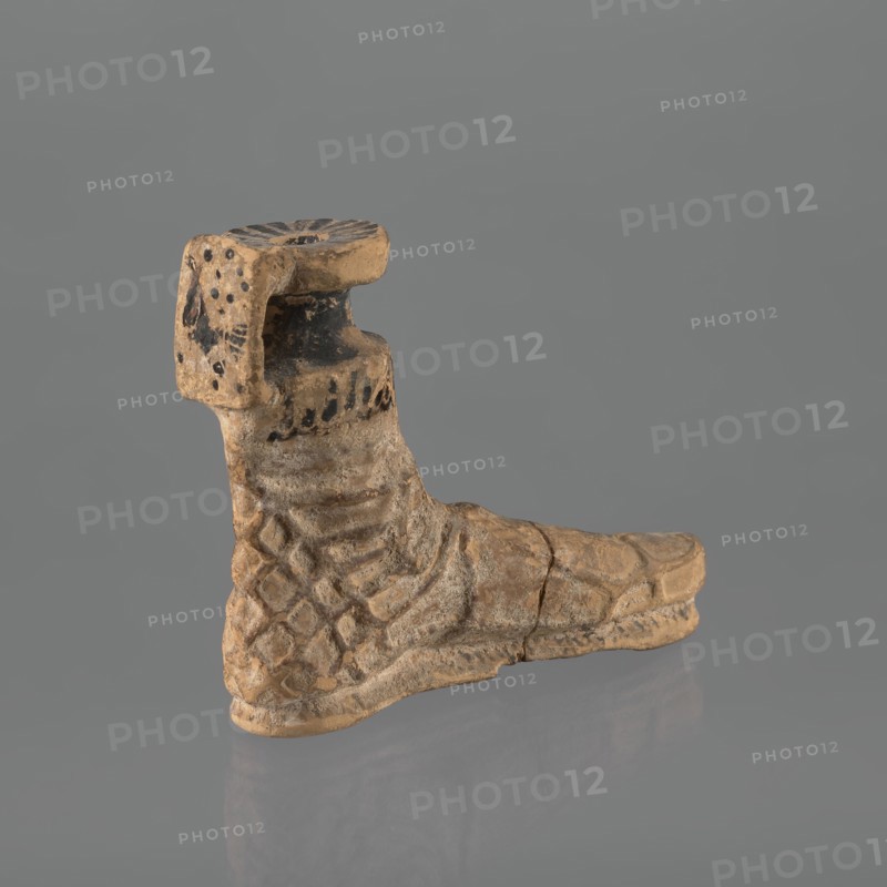 Foot-shaped aryballos, aryballos, antique vessel. Yellow-orange clay with mica; matte black engobe on the surface. Shaped as a foot shod in a sandal with a thick sole connected to braided leather straps. The foot ends above the ankle and merges into a narrow neck terminating in an outward-turned rim, decorated from above with radiating lines. A wide, ribbon-like handle is attached vertically to the spout, bearing an image of a rooster surrounded by dots. Preservation condition: the front part of the foot is filled with plaster. End of the 6th–beginning of the 5th century BC. Ionia, Turkey/Greece., Ionian workshop., A worn sculpted object resembling a boot shape sits against a plain gray background, showing a textured surface with carved patterns and a few incised lines. The piece displays a mottled palette of beige, tan, and light brown with darker brown and black smudges and spots in several areas, especially near the upper section. The surface appears rough and weathered with visible...