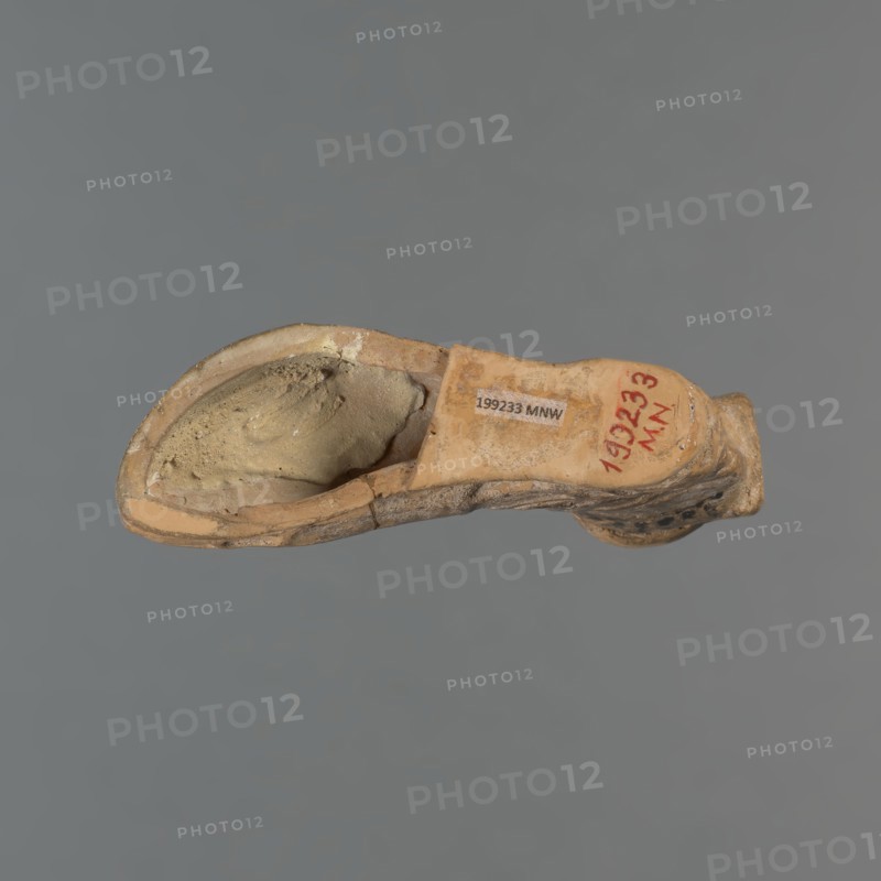 Foot-shaped aryballos, aryballos, antique vessel. Yellow-orange clay with mica; matte black engobe on the surface. Shaped like a foot shod in a sandal with a thick sole connected to braided leather straps. The foot ends above the ankle and merges into a narrow neck terminating in an outward-turned rim, decorated from above with radiating lines. A wide, ribbon-like handle is attached vertically to the spout, on which there is an image of a rooster surrounded by dots. Preservation condition: the front part of the foot is filled with plaster. End of the 6th–beginning of the 5th century BC. Made in an Ionian workshop; found in Ionia/Turkey/Greece., A worn, hollow object shaped like a curved vessel rests against a plain gray background; the object shows a beige and tan exterior with patches of darker brown and lighter cream tones, a rough textured inner cavity filled with a sandy, muted yellow-beige material, and areas of surface abrasion and small chips along the rim and edges.