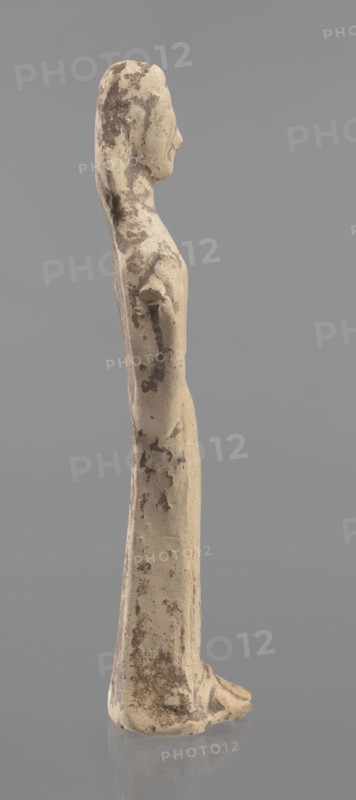 Terracotta figurine of a woman, unknown Boeotian workshop. Female mold-pressed figurine standing in a long chiton. Head set on a thick neck; oval face with a large nose and almond-shaped eyes. Arms bent at the elbows, forearms raised with hands pressed against the shoulders. Right hand holds a cylindrical object probably a fragment of a longer element. Traces of brown and red polychrome. Condition: left hand broken off, surface worn. Ca. 550 BC. Boeotia. Figurine; height 23 cm, width 7 cm., A slender standing figure viewed in profile, rendered in a weathered beige material with irregular darker brown patches and spotting across the surface; the figure has an elongated head with a pronounced nose, a long neck, and narrow shoulders leading into a straight, columnar torso and skirted lower body, with the suggestion of feet at the base; the surface shows rough texture, chips, and worn areas, and the background is a smooth mid-gray.