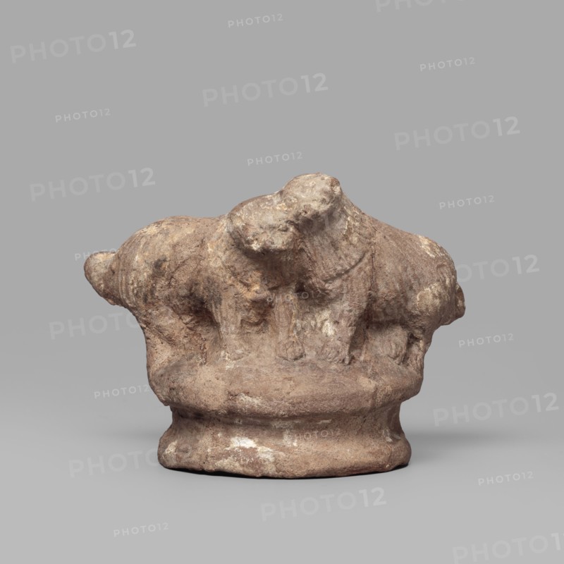 Two dogs, Two dogs shown schematically on a round, profiled base. The dogs are shown standing, with their heads touching. 332–30 BC; Ptolemaic Period, Tell Atrib; figurine; height 5.3 cm, width 7.1 cm., A small weathered clay sculpture depicting a group of animal-like forms atop a rounded base, with rough textured surfaces and visible wear; colors include shades of brown, tan, beige, and hints of off-white, set against a neutral gray background.
